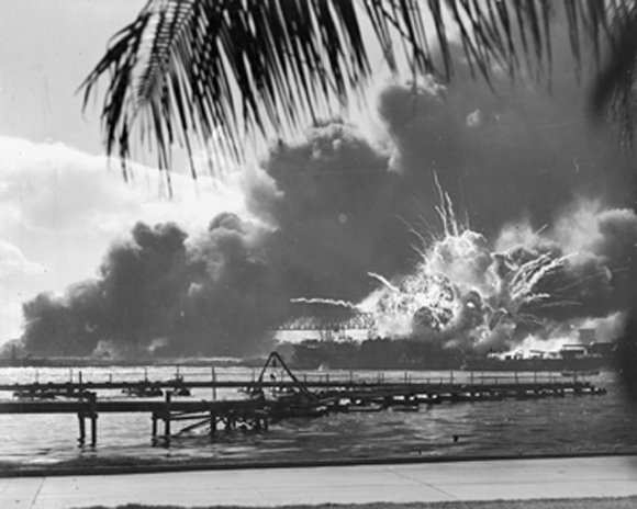 Photographic image of Pearl Harbor in smoke.  The image is taken at some distance and is framed by a palm tree and undamaged pavement.